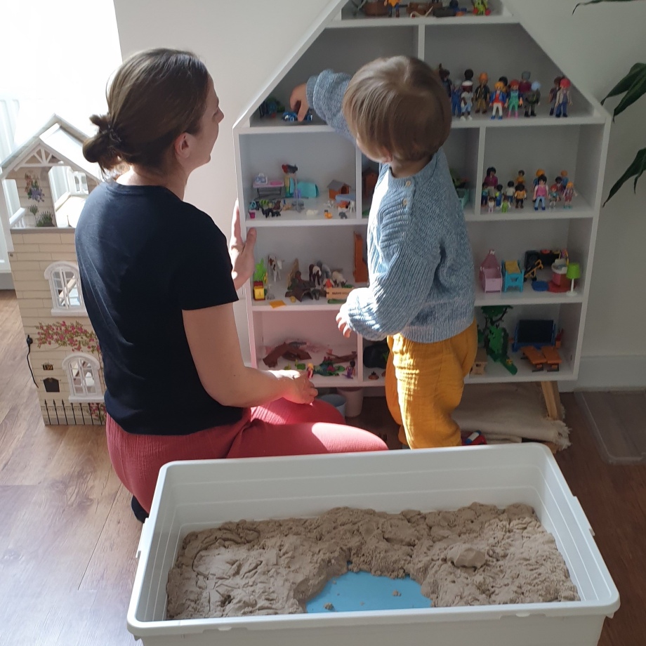 Psychologin bei der Arbeit mi einem Kleinkind, davor befindet sich eine weiße Kiste mit therapeutischem Sand und dahinter ein Regal mit Spielfiguren.